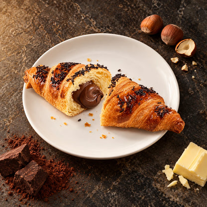 Croissant Chocolate y Avellana visto desde arriba en plato blanco, junto a avellanas, cacao, chocolate y mantequilla