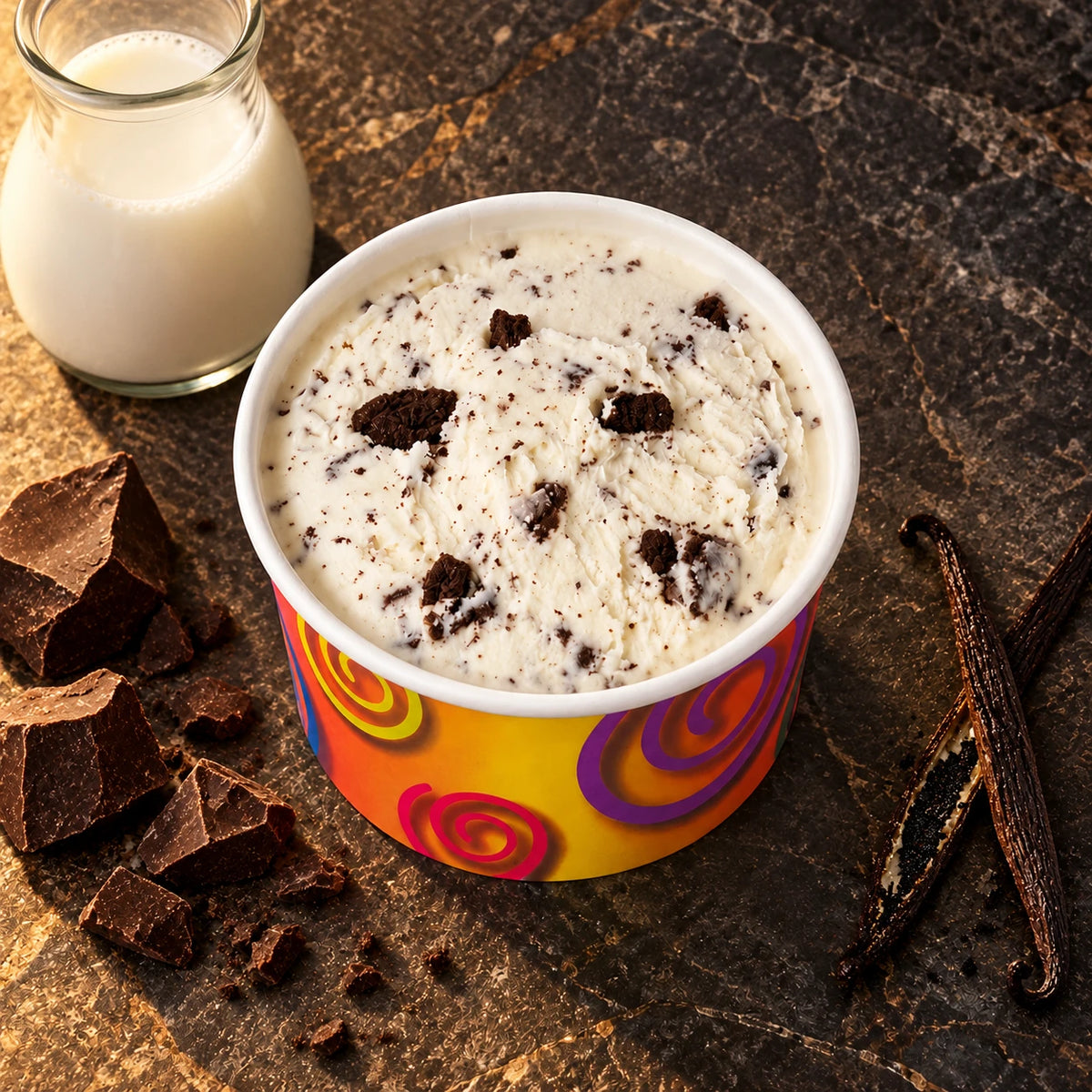 Vasito de helado cookies sobre piedra oscura con una botella de leche, trozos de chocolate y vainas de vainilla