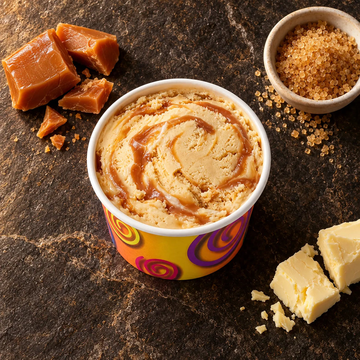 Vasito de helado de caramelo sobre piedra oscura con trozos de caramelo, azúcar moreno y mantequilla