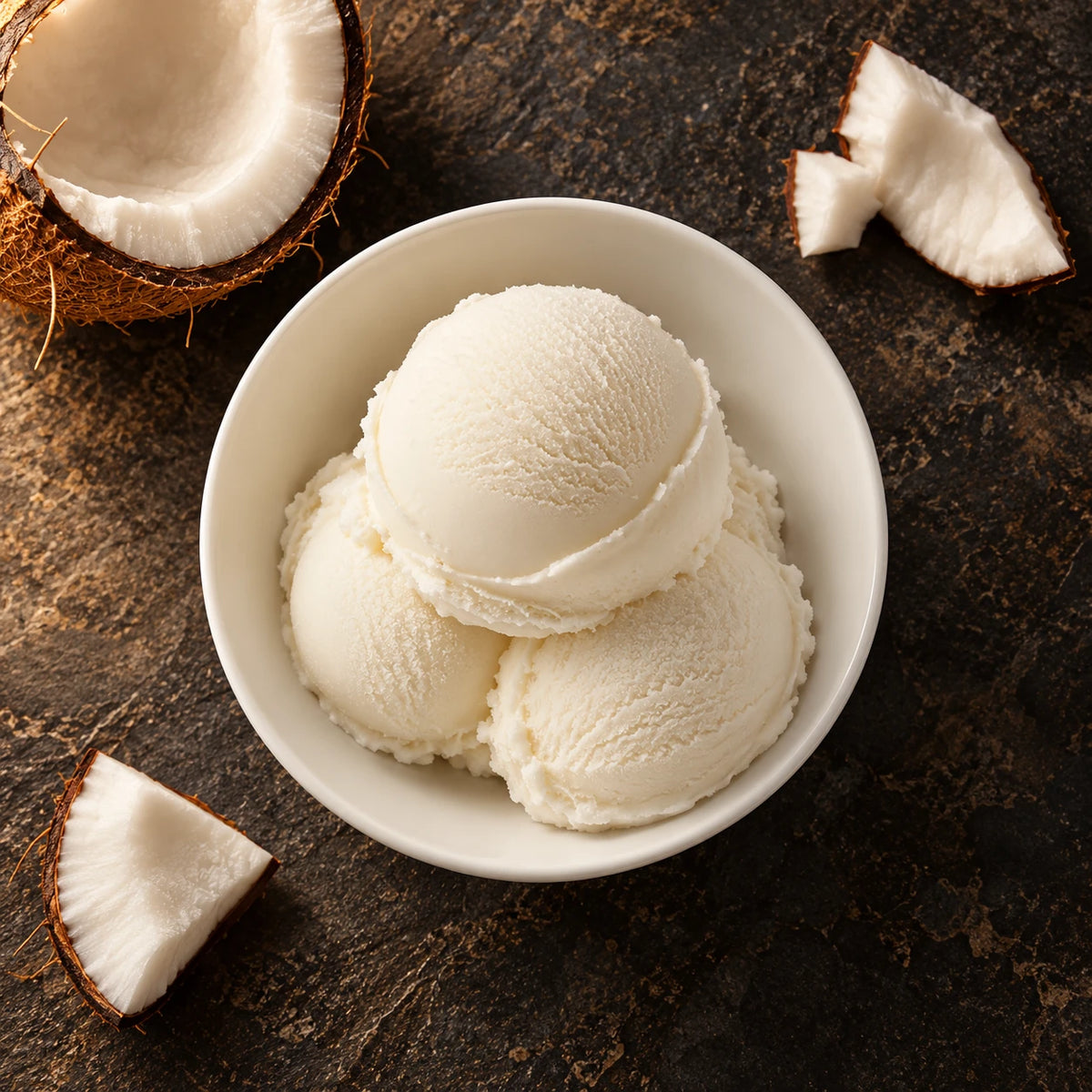 Tres bolas de helado de coco desde arriba en un cuenco blanco sobre piedra oscura, junto a trozos de coco