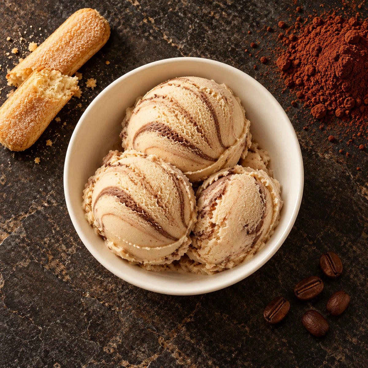 Tres bolas de helado de tiramisú desde arriba en un cuenco blanco sobre piedra oscura, con bizcochos de soletilla, cacao en polvo y granos de café