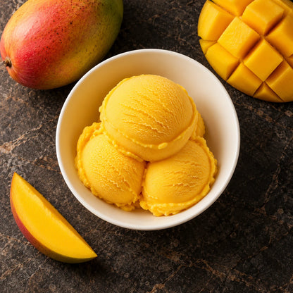 Tres bolas de sorbete de mango desde arriba en un cuenco blanco sobre piedra oscura, junto a un mango entero, una tajada y mango en dados