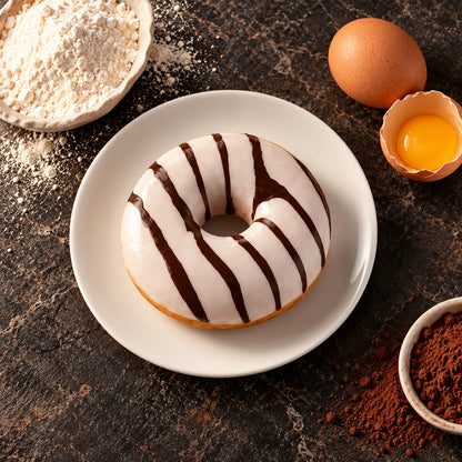 Donut w polewie waniliowej z góry na białym talerzu, obok mąka, jajko i kakao